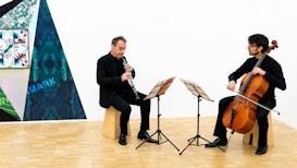 Two musicians in black attire are playing instruments in a spacious, well-lit room. One is playing the clarinet while the other plays the cello. They are seated on wooden stools with music stands in front of them. The background features a modern art piece with geometric patterns and vibrant colors.