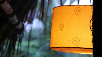 A warm, glowing lampshade with spiral patterns hangs, casting a soft light. The background features blurred leaves and a structure resembling a thatched roof, suggesting a cozy and natural setting.