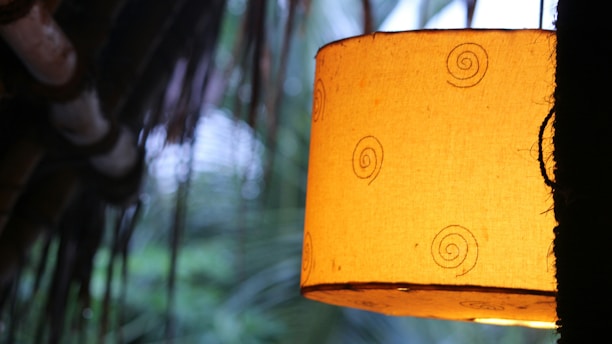 A warm, glowing lampshade with spiral patterns hangs, casting a soft light. The background features blurred leaves and a structure resembling a thatched roof, suggesting a cozy and natural setting.