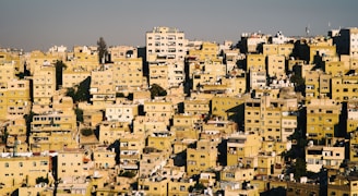 A densely packed urban landscape featuring numerous rectangular buildings, predominantly in shades of yellow and beige. The structures are closely arranged on a sloping hillside, with various architectural styles and heights. Many buildings appear residential, with visible windows, balconies, and rooftops dotted with satellite dishes and water tanks.