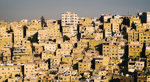 A densely packed urban landscape featuring numerous rectangular buildings, predominantly in shades of yellow and beige. The structures are closely arranged on a sloping hillside, with various architectural styles and heights. Many buildings appear residential, with visible windows, balconies, and rooftops dotted with satellite dishes and water tanks.