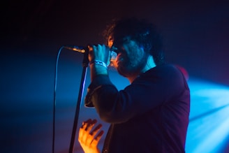 A person passionately singing into a microphone on stage, illuminated by moody blue and orange lighting, creating a dramatic contrast. The background is dark with a spotlight adding depth and atmosphere to the scene.