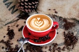 A red cup filled with a beautifully crafted latte, featuring intricate latte art in the shape of a heart and leaves. The cup rests on a matching saucer placed on a burlap sack. Coffee beans and ground coffee are artfully scattered around the cup. A large pinecone and two spoons are also visible in the composition.