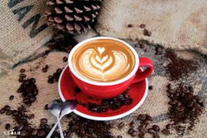 A red cup filled with a beautifully crafted latte, featuring intricate latte art in the shape of a heart and leaves. The cup rests on a matching saucer placed on a burlap sack. Coffee beans and ground coffee are artfully scattered around the cup. A large pinecone and two spoons are also visible in the composition.