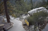 A winding concrete path leads through a forested mountainous area, surrounded by tall trees and large boulders. A wooden railing lines one side of the path, offering safety and guiding visitors. A large rock is prominently positioned beside the path, with small shrubs and vegetation scattered throughout the scene. Two people are visible; one sitting on a bench and another walking along the path. A yellow sign is visible, suggesting the location may be a natural park or trail.