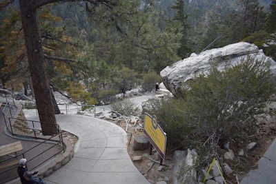 A winding concrete path leads through a forested mountainous area, surrounded by tall trees and large boulders. A wooden railing lines one side of the path, offering safety and guiding visitors. A large rock is prominently positioned beside the path, with small shrubs and vegetation scattered throughout the scene. Two people are visible; one sitting on a bench and another walking along the path. A yellow sign is visible, suggesting the location may be a natural park or trail.