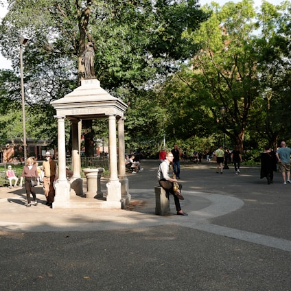 A park scene with a stone monument labeled 'Charity' at the center, surrounded by lush greenery and several people walking or sitting. There are benches and a paved path, with sunlight filtering through the trees.