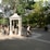 A park scene with a stone monument labeled 'Charity' at the center, surrounded by lush greenery and several people walking or sitting. There are benches and a paved path, with sunlight filtering through the trees.