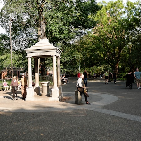A park scene with a stone monument labeled 'Charity' at the center, surrounded by lush greenery and several people walking or sitting. There are benches and a paved path, with sunlight filtering through the trees.