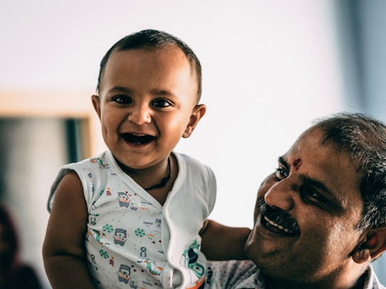 A smiling baby is being held by an adult, who is also smiling. The baby is wearing a white shirt with cartoon prints, and the adult has a tilaka mark on the forehead. The background is unfocused with a soft, natural light.