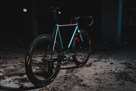 A sleek, modern bicycle is positioned in an industrial, dimly lit setting. The bike features a minimalist design, with a focus on its smooth, aerodynamic frame and thick tires. The area is scattered with debris, and the overall ambiance is dark and gritty, creating a contrast with the bike's shinier, colorful highlights.