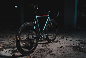 A sleek, modern bicycle is positioned in an industrial, dimly lit setting. The bike features a minimalist design, with a focus on its smooth, aerodynamic frame and thick tires. The area is scattered with debris, and the overall ambiance is dark and gritty, creating a contrast with the bike's shinier, colorful highlights.