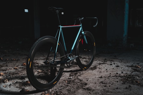 A sleek, modern bicycle is positioned in an industrial, dimly lit setting. The bike features a minimalist design, with a focus on its smooth, aerodynamic frame and thick tires. The area is scattered with debris, and the overall ambiance is dark and gritty, creating a contrast with the bike's shinier, colorful highlights.