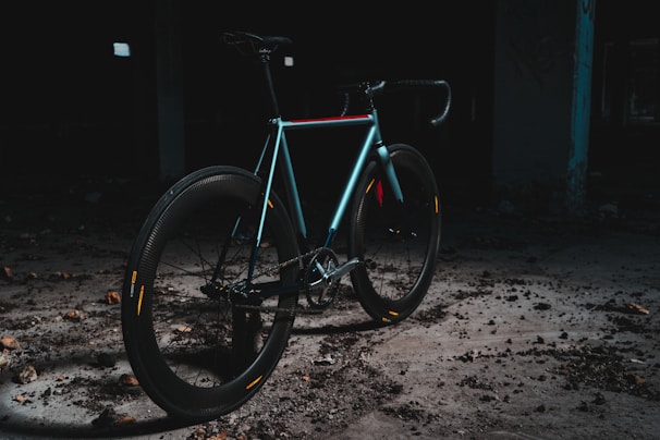A sleek, modern bicycle is positioned in an industrial, dimly lit setting. The bike features a minimalist design, with a focus on its smooth, aerodynamic frame and thick tires. The area is scattered with debris, and the overall ambiance is dark and gritty, creating a contrast with the bike's shinier, colorful highlights.