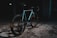 A sleek, modern bicycle is positioned in an industrial, dimly lit setting. The bike features a minimalist design, with a focus on its smooth, aerodynamic frame and thick tires. The area is scattered with debris, and the overall ambiance is dark and gritty, creating a contrast with the bike's shinier, colorful highlights.