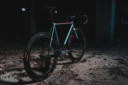 A sleek, modern bicycle is positioned in an industrial, dimly lit setting. The bike features a minimalist design, with a focus on its smooth, aerodynamic frame and thick tires. The area is scattered with debris, and the overall ambiance is dark and gritty, creating a contrast with the bike's shinier, colorful highlights.
