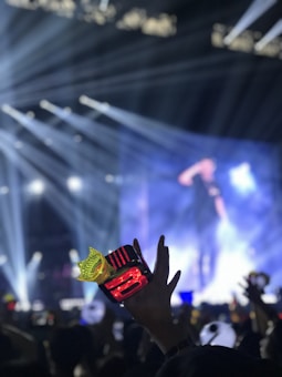 A concert scene with a crowd, featuring a hand holding a red and yellow lightstick prominently in the foreground. Bright stage lights and beams create a dynamic and lively atmosphere, while a large screen displays a blurry image of a person on stage.