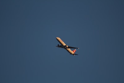 A commercial airplane is flying across a clear blue sky with a visible logo on the tail fin. The aircraft is illuminated by natural light, suggesting it might be flying during the day.