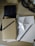 A collection of craft materials arranged on a light wooden surface, including brown cards with a label, a black pencil, white paper, and a three-dimensional white star with black speckles. A black, beaded necklace and a brown paper bag are also visible nearby.