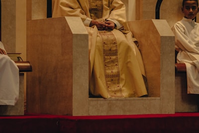 A central figure wearing ornate golden robes sits on a large, stone-like chair. The chair is set on a red carpet, and two individuals in white robes are seated on either side, slightly lower.