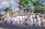 A group of people in traditional attire are participating in a cultural parade. They are dressed in white shirts and checkered sarongs, some holding musical instruments. The background shows tropical plants and a stone wall, suggesting a tropical location.