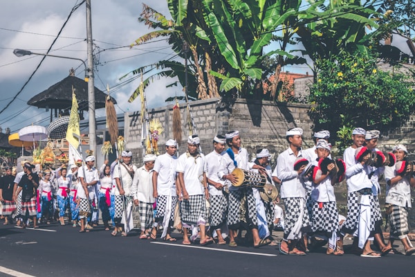 A group of people in traditional attire are participating in a cultural parade. They are dressed in white shirts and checkered sarongs, some holding musical instruments. The background shows tropical plants and a stone wall, suggesting a tropical location.