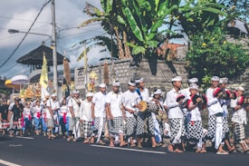 A group of people in traditional attire are participating in a cultural parade. They are dressed in white shirts and checkered sarongs, some holding musical instruments. The background shows tropical plants and a stone wall, suggesting a tropical location.
