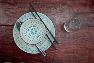 A rustic wooden table serves as the backdrop for a set of intricately patterned ceramic plates in teal and white. Resting on the plates are black chopsticks and a shiny spoon, while a clear glass filled with water stands to the side.