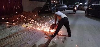 A person is using a power tool to cut through metal rods at night on the side of a road. Sparks are flying from the tool as it makes contact with the metal. There are other vehicles and people visible in the background, suggesting an urban environment.
