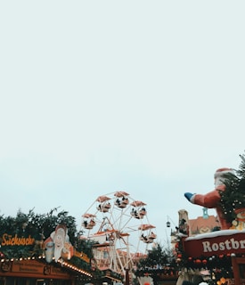 A festive Christmas market scene with a Ferris wheel in the background and a large Santa Claus figure on the right. Stalls decorated with wreaths and festive lights are visible, with patrons milling around. The overall atmosphere is cheerful and holiday-themed.