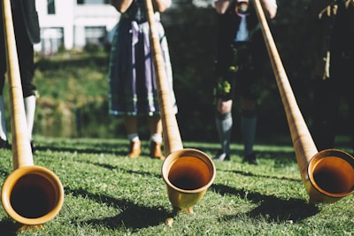 A group of people wearing traditional Bavarian attire are playing long wooden alphorns on a grassy area. Sunlight gently illuminates the scene, casting soft shadows on the grass. The background includes a white building and foliage.