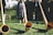 A group of people wearing traditional Bavarian attire are playing long wooden alphorns on a grassy area. Sunlight gently illuminates the scene, casting soft shadows on the grass. The background includes a white building and foliage.