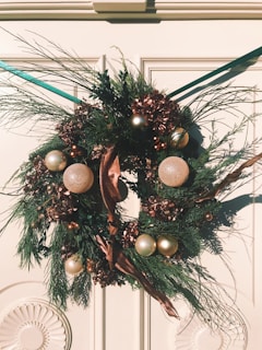 A decorative wreath hangs on a door, composed of pine branches, metallic ornaments, and pinecones. The ornaments are gold and silver, with a mix of natural and festive elements. A brown ribbon interweaves through the wreath, adding to the holiday theme. The door behind the wreath has a subtle ornate design.
