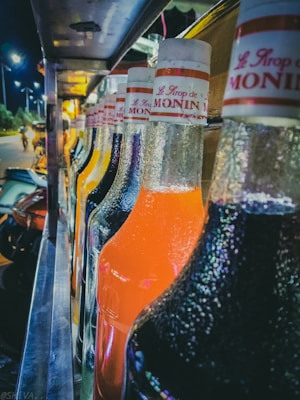 A row of colorful syrup bottles is lined up, with condensation visible on their surfaces. Each bottle is labeled 'Le Sirop de MONIN' with various vibrant shades, including orange and deep red. The scene takes place outdoors, with streetlights illuminating the background and a blurred figure passing by.