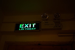 A green illuminated exit sign with Vietnamese text 'Lối Thoát' is mounted on a wall. Above the sign are two emergency lights. The wall and ceiling appear beige and the lighting creates a dim atmosphere.
