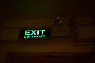 A green illuminated exit sign with Vietnamese text 'Lối Thoát' is mounted on a wall. Above the sign are two emergency lights. The wall and ceiling appear beige and the lighting creates a dim atmosphere.