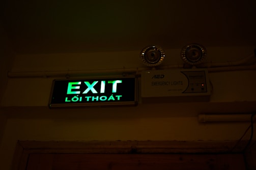A green illuminated exit sign with Vietnamese text 'Lối Thoát' is mounted on a wall. Above the sign are two emergency lights. The wall and ceiling appear beige and the lighting creates a dim atmosphere.