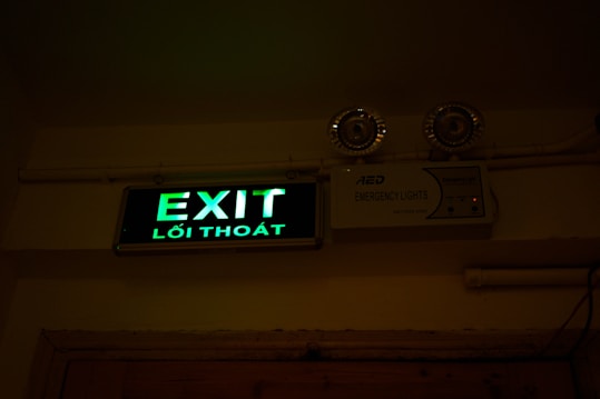 A green illuminated exit sign with Vietnamese text 'Lối Tho&aacute;t' is mounted on a wall. Above the sign are two emergency lights. The wall and ceiling appear beige and the lighting creates a dim atmosphere.