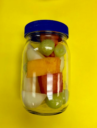 A clear glass jar with a blue lid contains colorful fruit pieces, including green grapes, watermelon, and other assorted fruits, placed against a vivid yellow background.