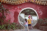 Three children play near a traditional circular entrance, surrounded by a red wall with greenery. One child wears a balloon hat, while the other two are dressed in casual clothes.