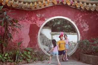 Three children play near a traditional circular entrance, surrounded by a red wall with greenery. One child wears a balloon hat, while the other two are dressed in casual clothes.