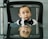 A young child dressed in a white shirt and black bow tie sits on a black mesh office chair. The child has a curious expression and is reflected on a glass table in front. In the background, an office-like setting is partially visible with a chair and a camera on a stand.