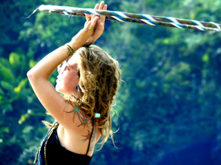 A woman with wavy blonde hair is seen in profile, raising her arms above her head, holding a twisted, multicolored hula hoop. She wears glasses and various bracelets, and has a serene expression. The background is a lush, green forest, slightly blurred.