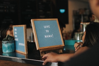 Two wooden-framed message boards are displayed on a countertop. One board contains the message 'LOVE YO MAMA!' and the other has partial text visible. A patterned blue plant pot with a small succulent is on the counter beside the boards. People are in close proximity, one person is leaning on the counter, suggesting a relaxed atmosphere in what appears to be a cafe or similar setting.