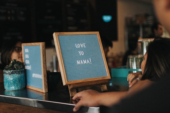 Two wooden-framed message boards are displayed on a countertop. One board contains the message 'LOVE YO MAMA!' and the other has partial text visible. A patterned blue plant pot with a small succulent is on the counter beside the boards. People are in close proximity, one person is leaning on the counter, suggesting a relaxed atmosphere in what appears to be a cafe or similar setting.