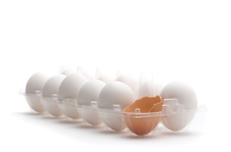 A row of white eggs is arranged in a transparent plastic egg carton, with one brown eggshell cracked open among them.