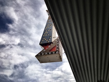 A large, metallic sign with an interesting geometric design featuring several round bulbs is seen against a backdrop of a cloudy sky. The sign is positioned vertically and appears to be attached to a corrugated metal structure extending diagonally into the frame.