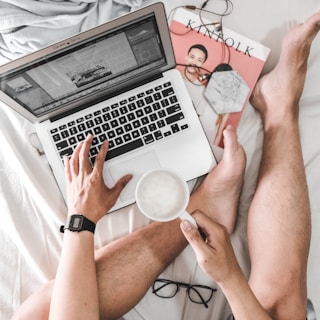 A person is sitting on a bed with a laptop on their lap. They are using one hand to navigate the computer while holding a white mug in the other. Nearby, a pair of glasses, earphones, and a magazine with the title 'Kinfolk' lie next to them. The setup suggests a relaxed, work-from-home or leisure reading environment.