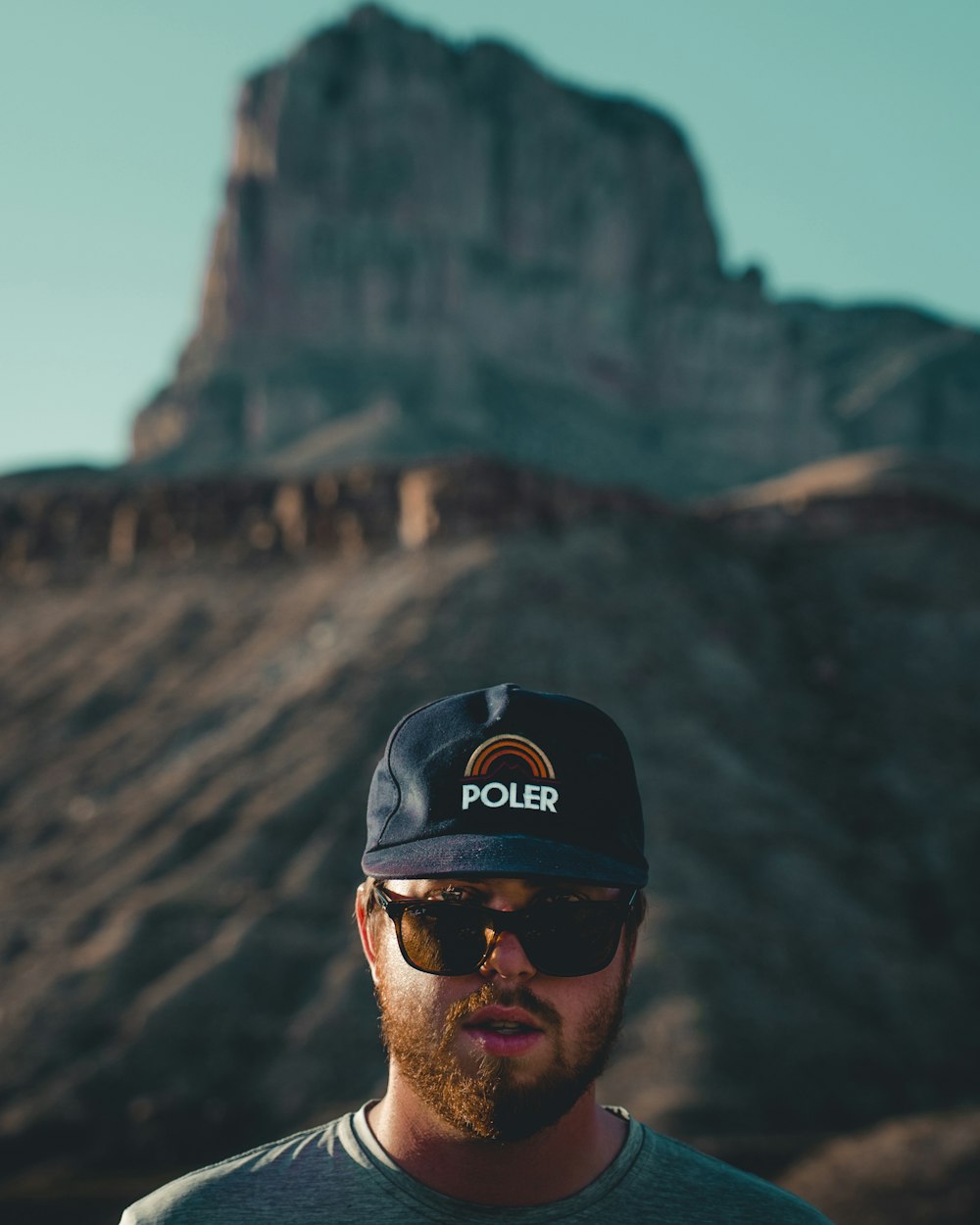 man in grey shirt wearing black Poler cap standing with rock canyon on background