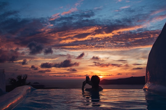 A person is immersed in a pool, holding a camera or phone towards a dramatic sunset. The sky is filled with vibrant hues of orange, pink, and blue, with scattered clouds enhancing the visual effect. The silhouette of the individual adds to the serene and picturesque quality of the scene.
